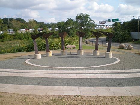 Outdoor sculpture with metal abstract figures arranged in a circle on a gravel surface near a highway.