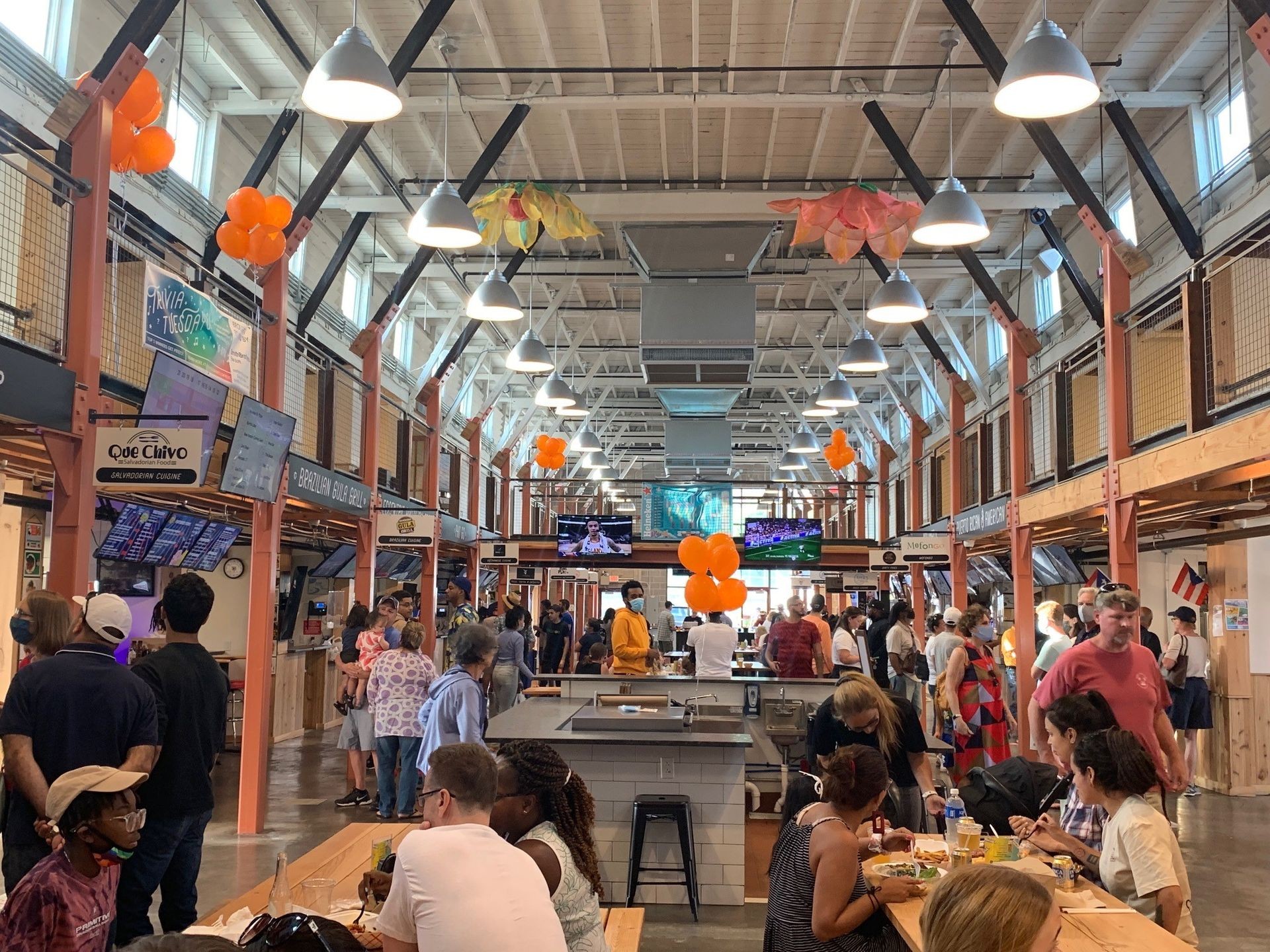 Crowded food hall with people dining at communal tables and various food vendor stalls under high ceilings.